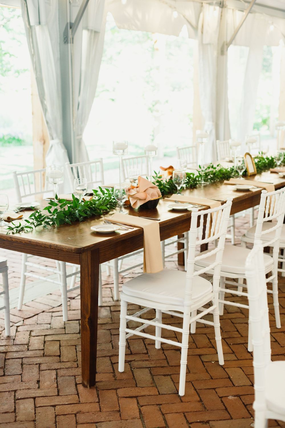 Elegant wedding reception setup with long wooden tables, white chairs and greenery runners under a tent.