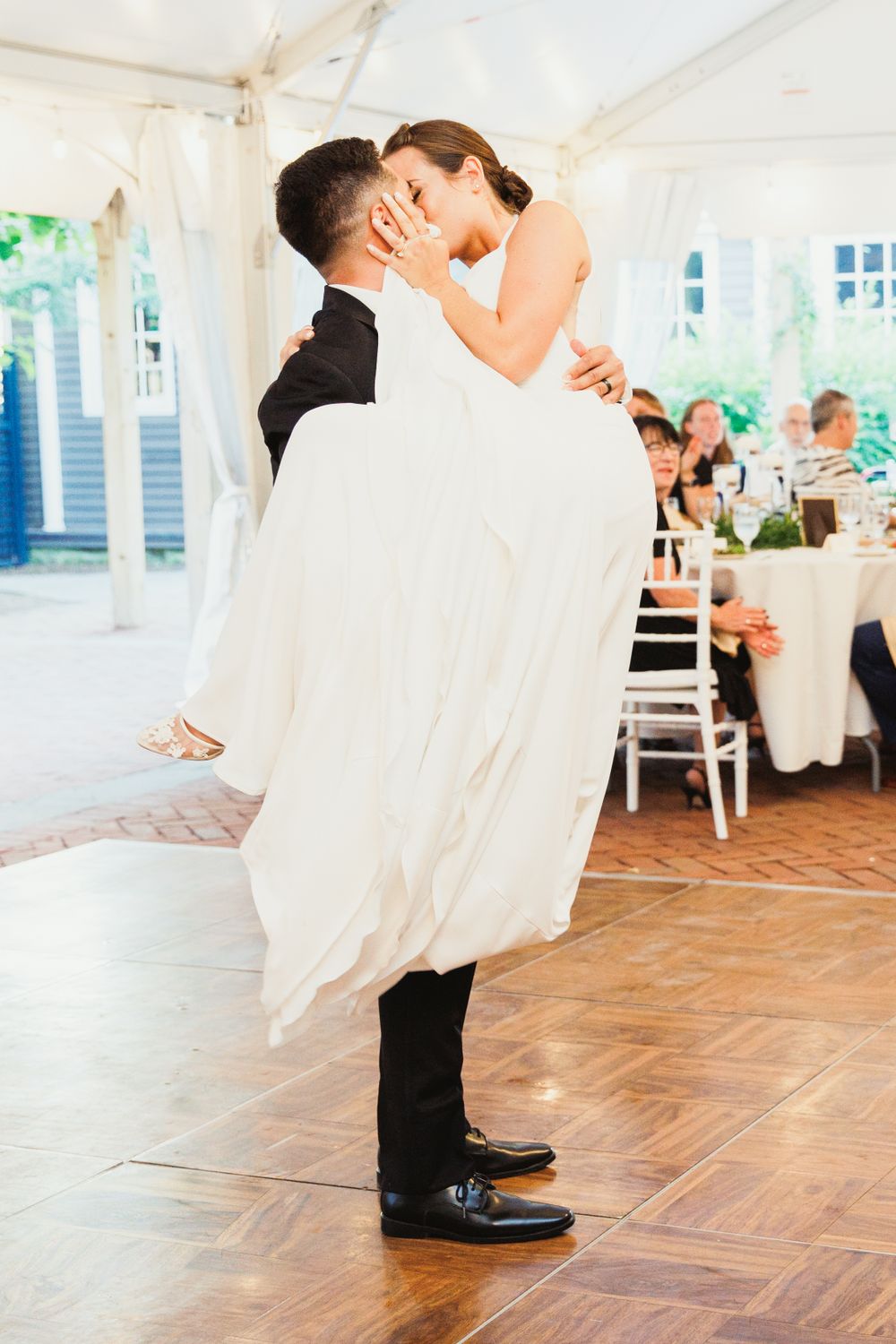 Groom lifts bride in flowing white dress during romantic dance moment at tented wedding reception.