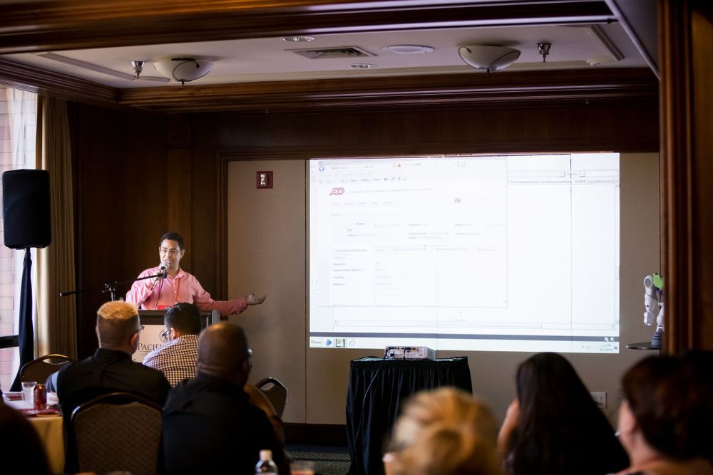 Conference presenter in pink shirt gestures during presentation with projected screen in background.