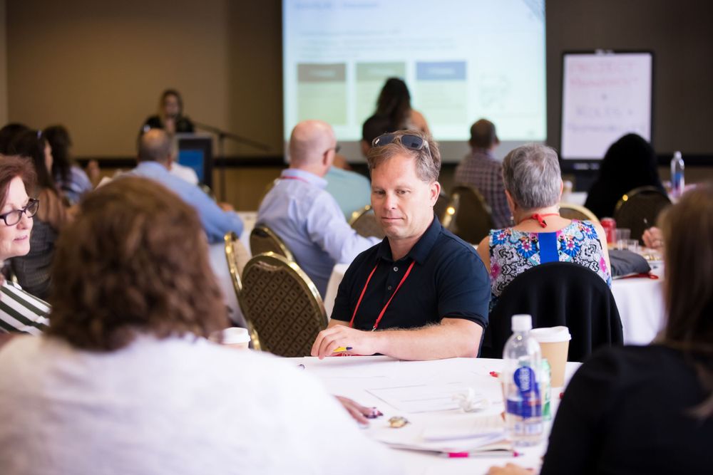 Conference attendees seated at round tables in a meeting room with a projection screen visible.