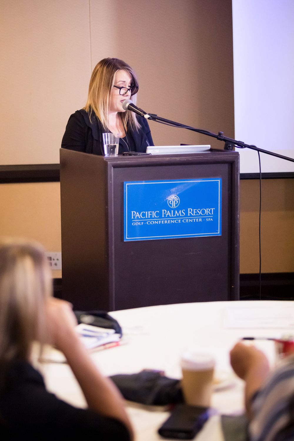 Conference presenter speaking at podium with audience members visible in foreground.
