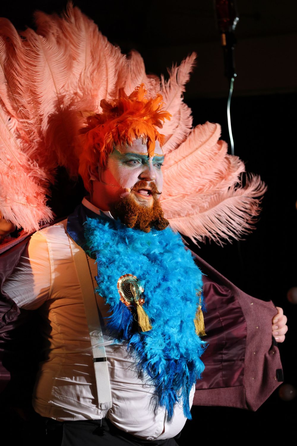 Performer in theatrical costume wearing pink feathers and bright blue makeup during a dramatic stage show.