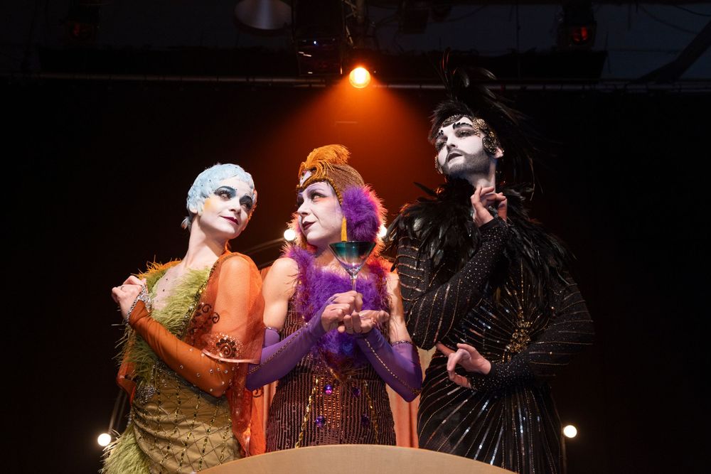 Three performers in elaborate costumes performing on stage under warm lighting.