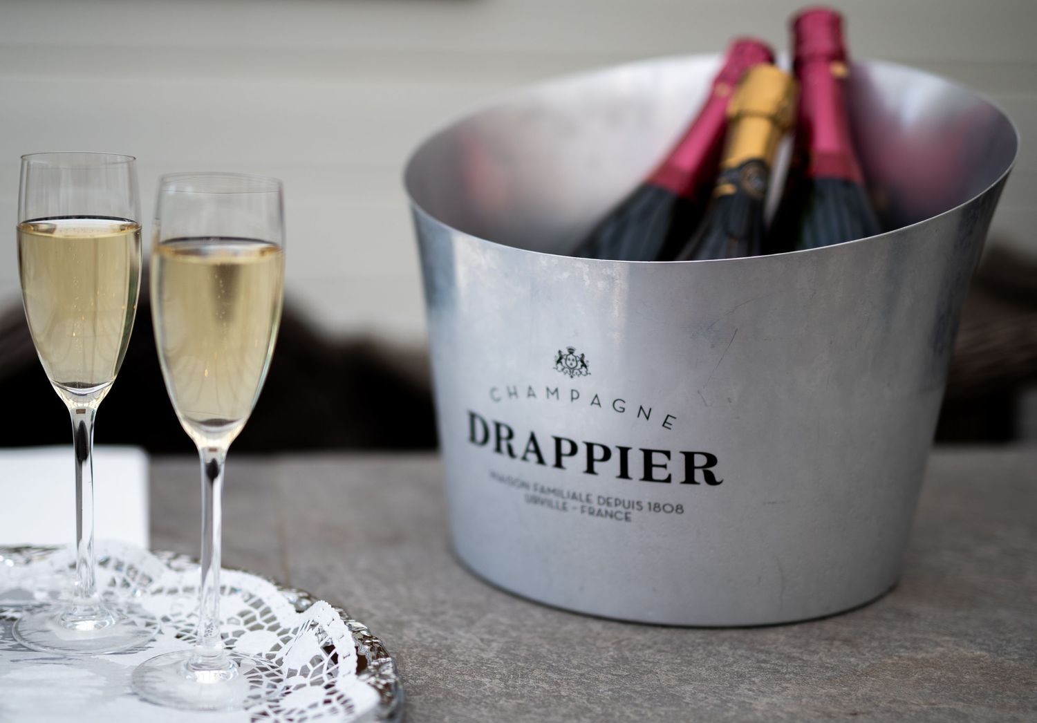 Two champagne flutes beside a Drappier Champagne ice bucket filled with bottles on marble surface.