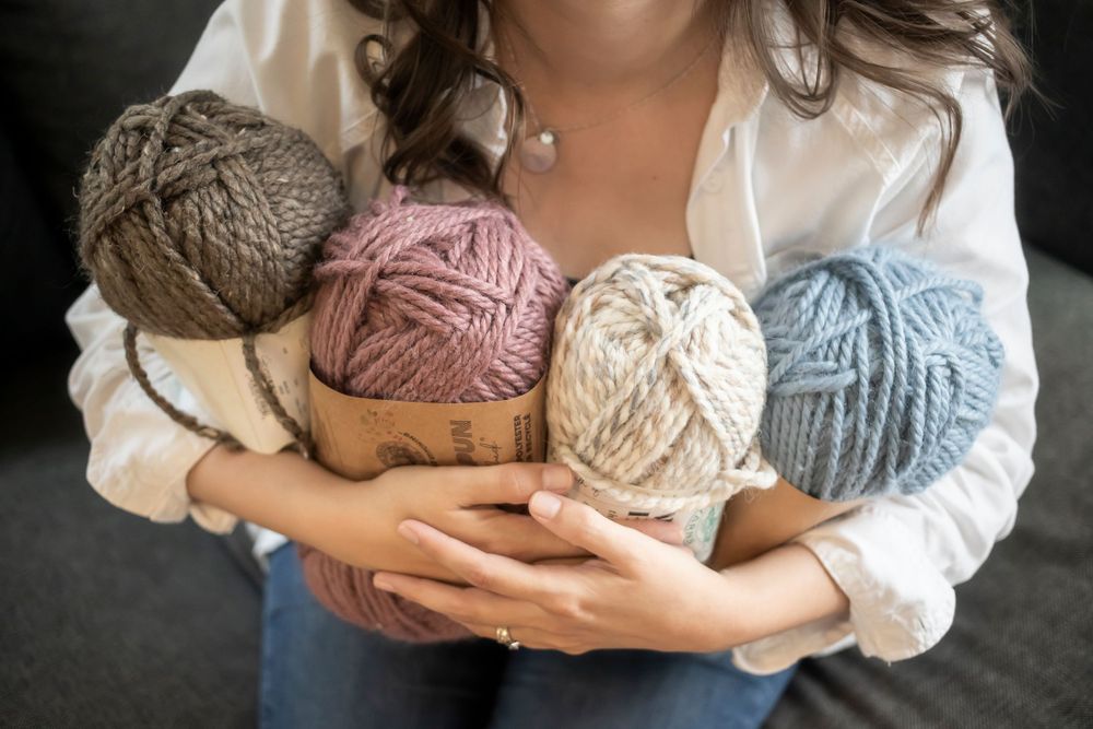 Hands holding collection of yarn balls in gray, pink, beige and blue colors.