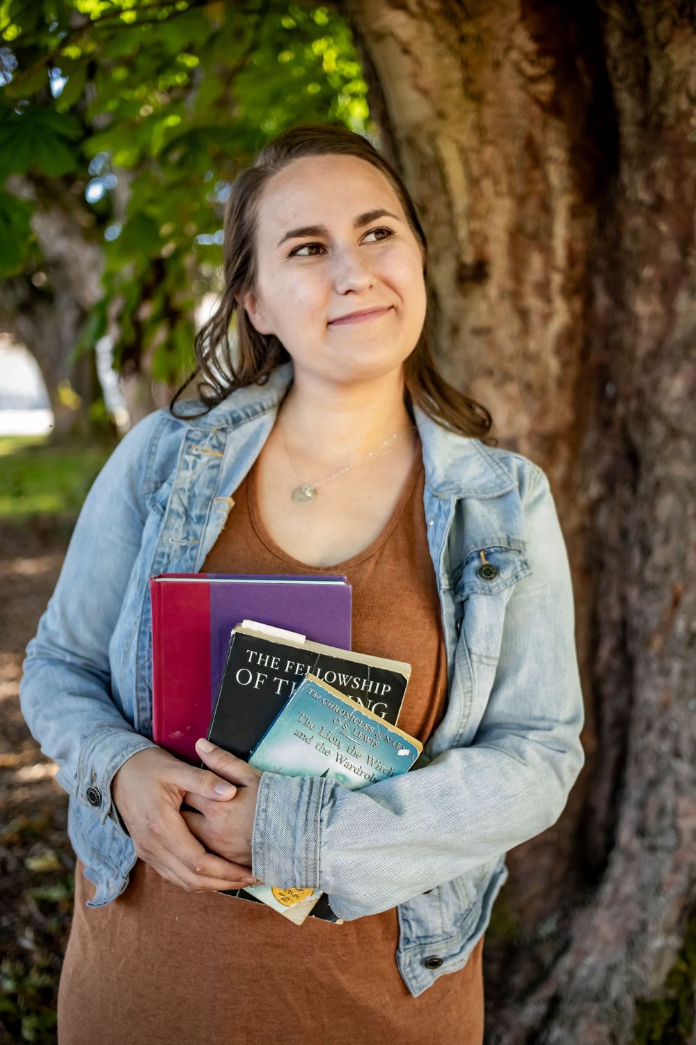 Person in denim jacket holding books against tree trunk background.