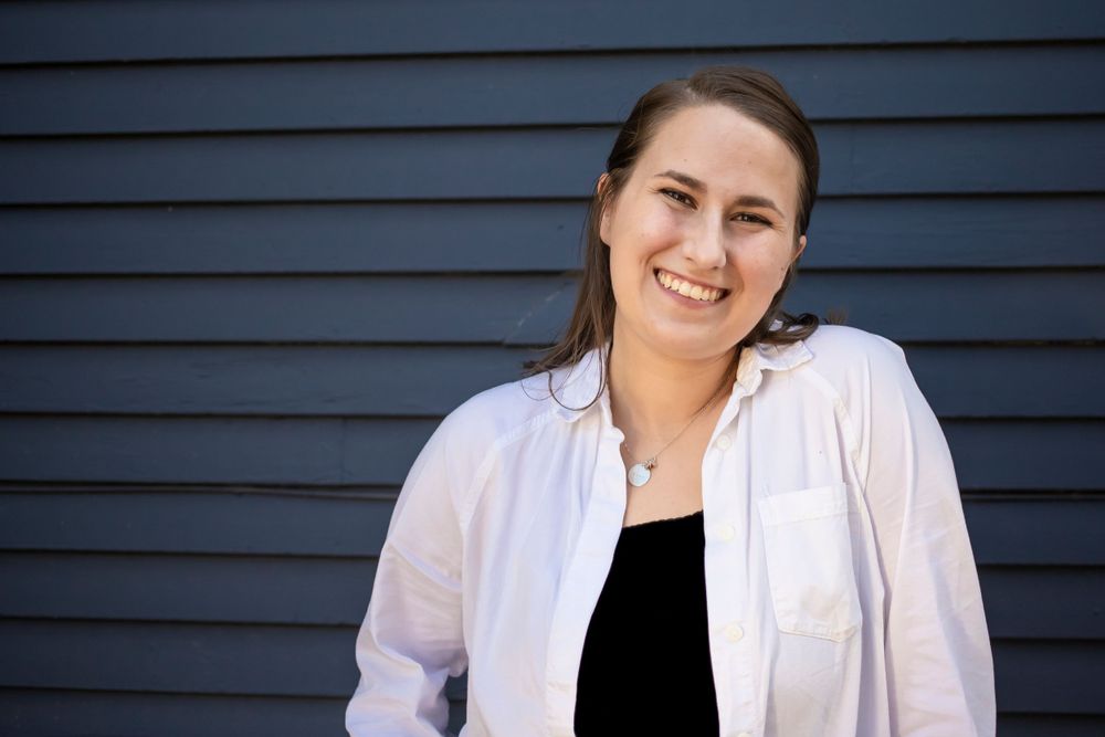 Portrait against blue siding wearing white blazer and black top.