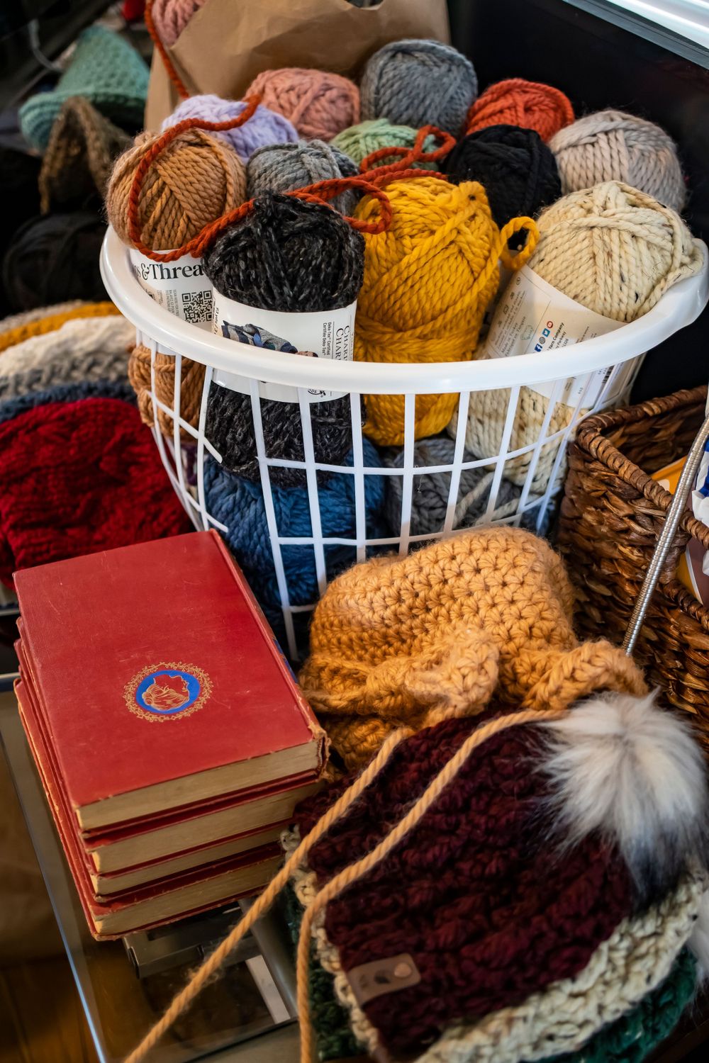 White basket filled with colorful yarn balls and knitting supplies next to red books.