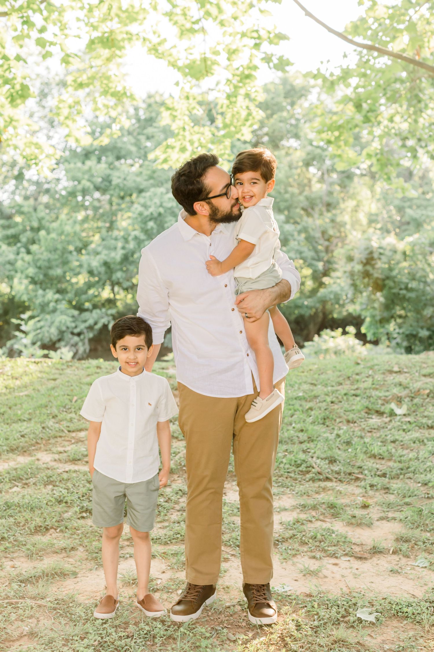 Three people share joyful moment together on grassy hillside wearing white shirts and khaki clothing.