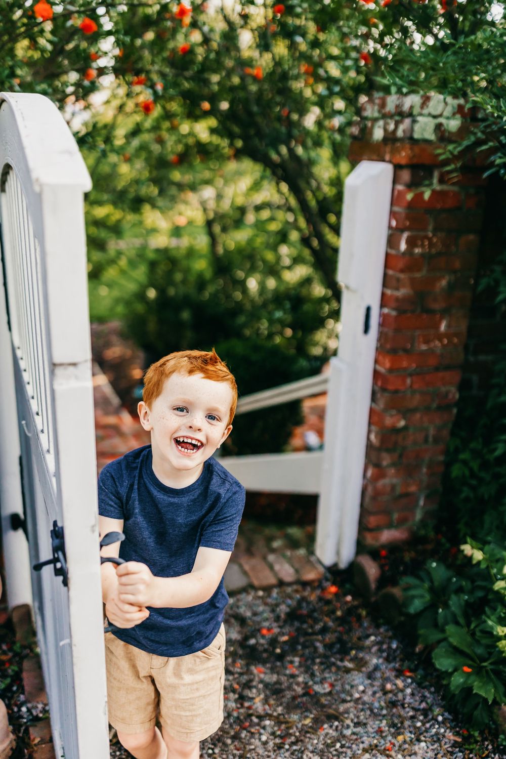Young child playing at white garden gate entrance with orange flowers and brick walls.