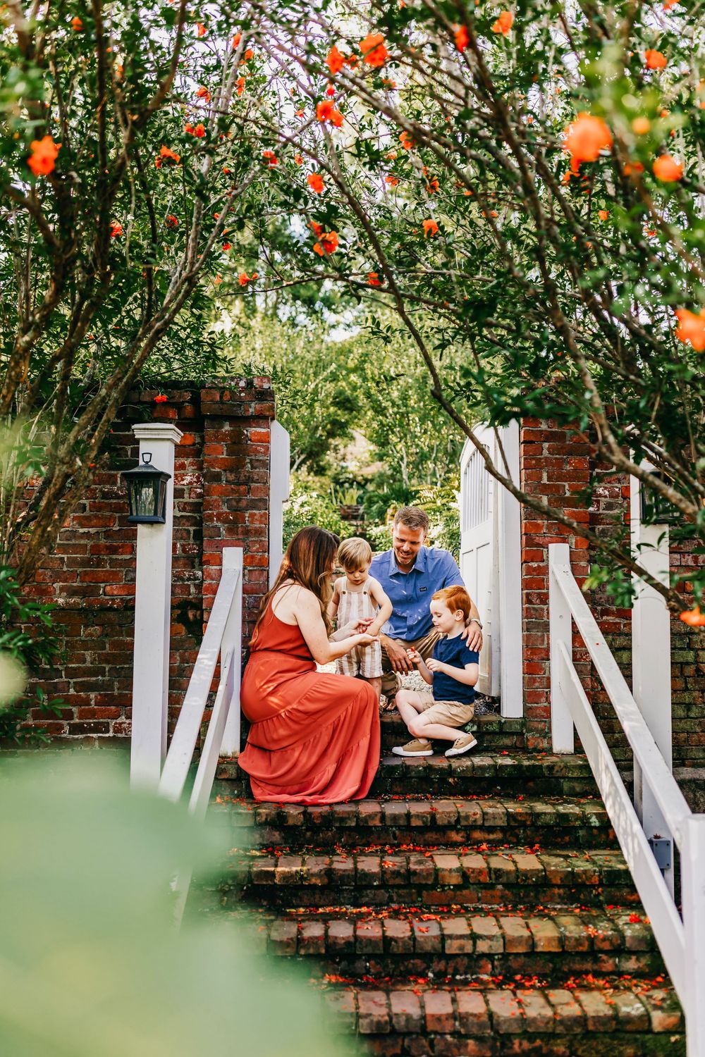 A tender family moment captured on brick garden steps surrounded by blooming orange flowers and white railings.