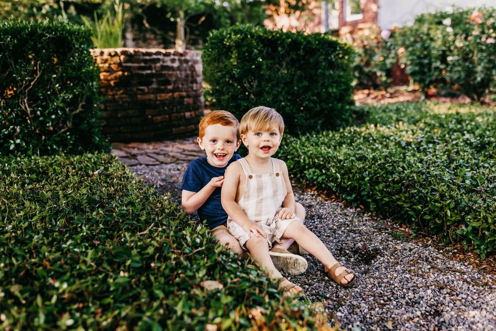 Two small children sit together on a gravel garden path surrounded by manicured hedges.