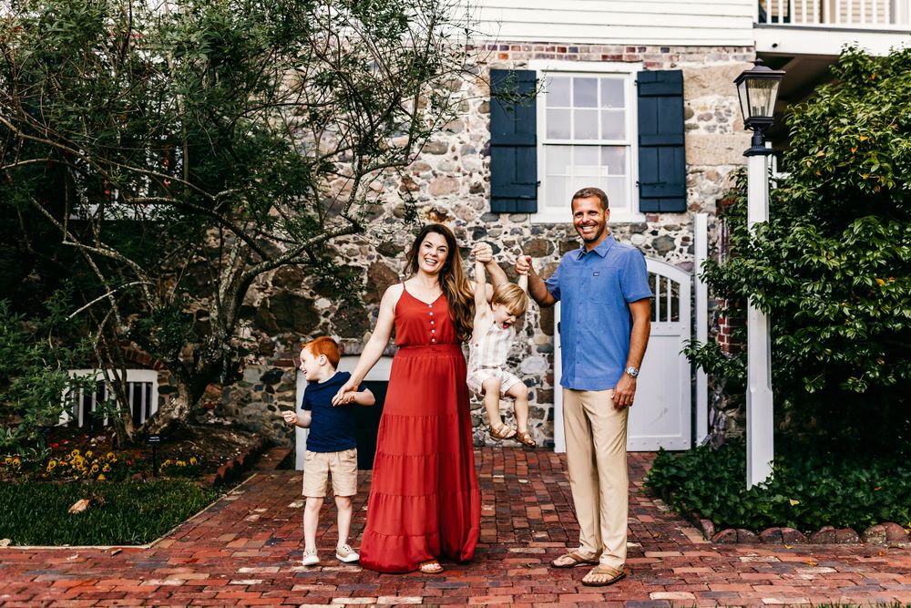 A family stands together on a brick path in front of a stone house with navy blue shutters and climbing vines.