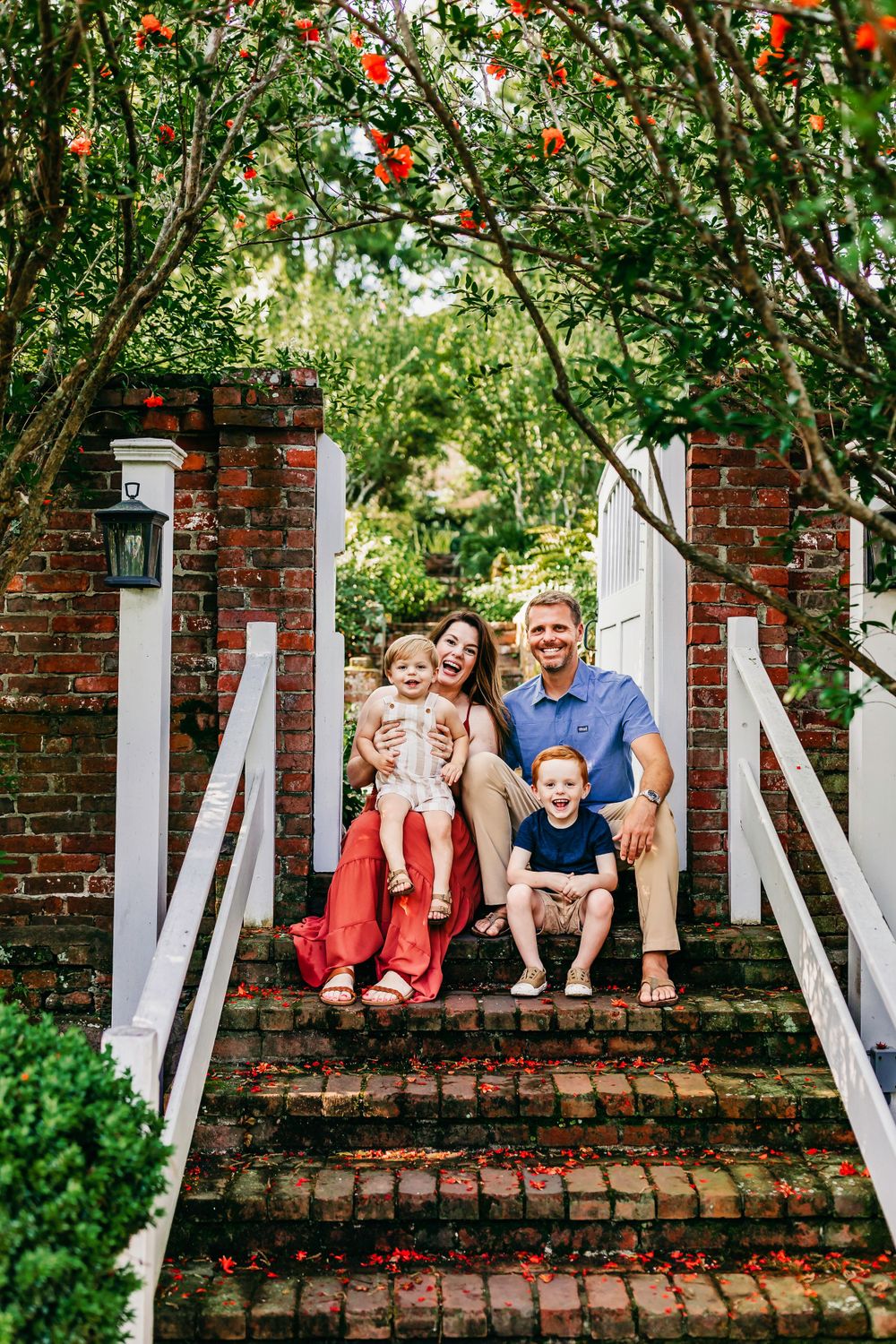 Family sitting together on brick steps with white railings and flowering trees overhead.