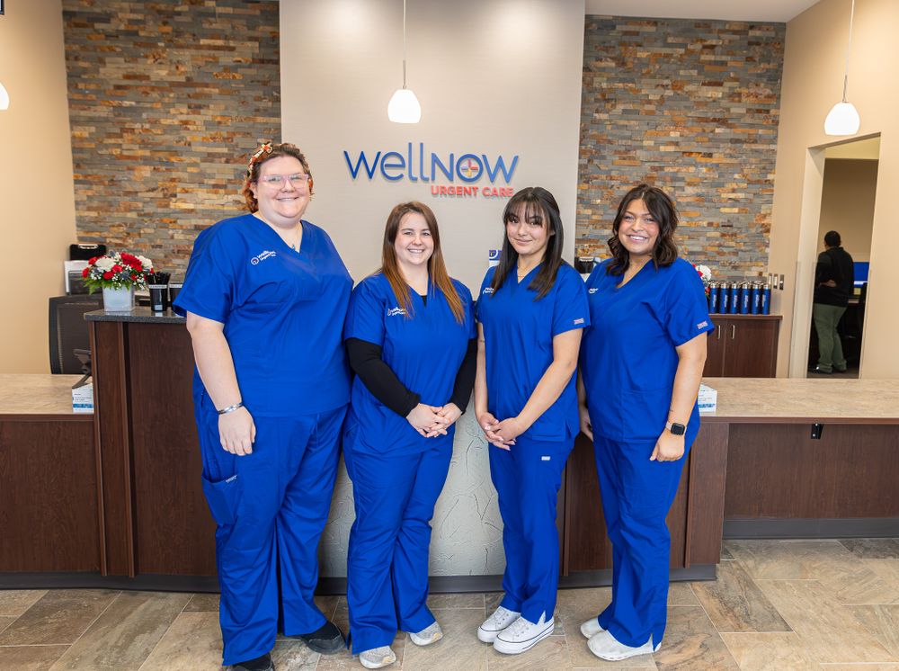 Healthcare workers in blue scrubs pose in front of a WellNow clinic reception desk.