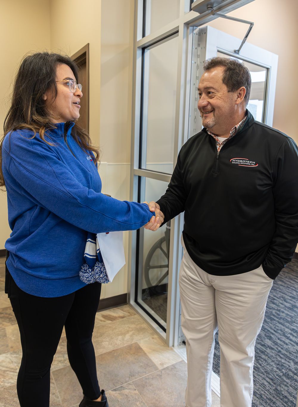 Professional greeting and handshake in an office hallway between colleagues in blue and black attire.