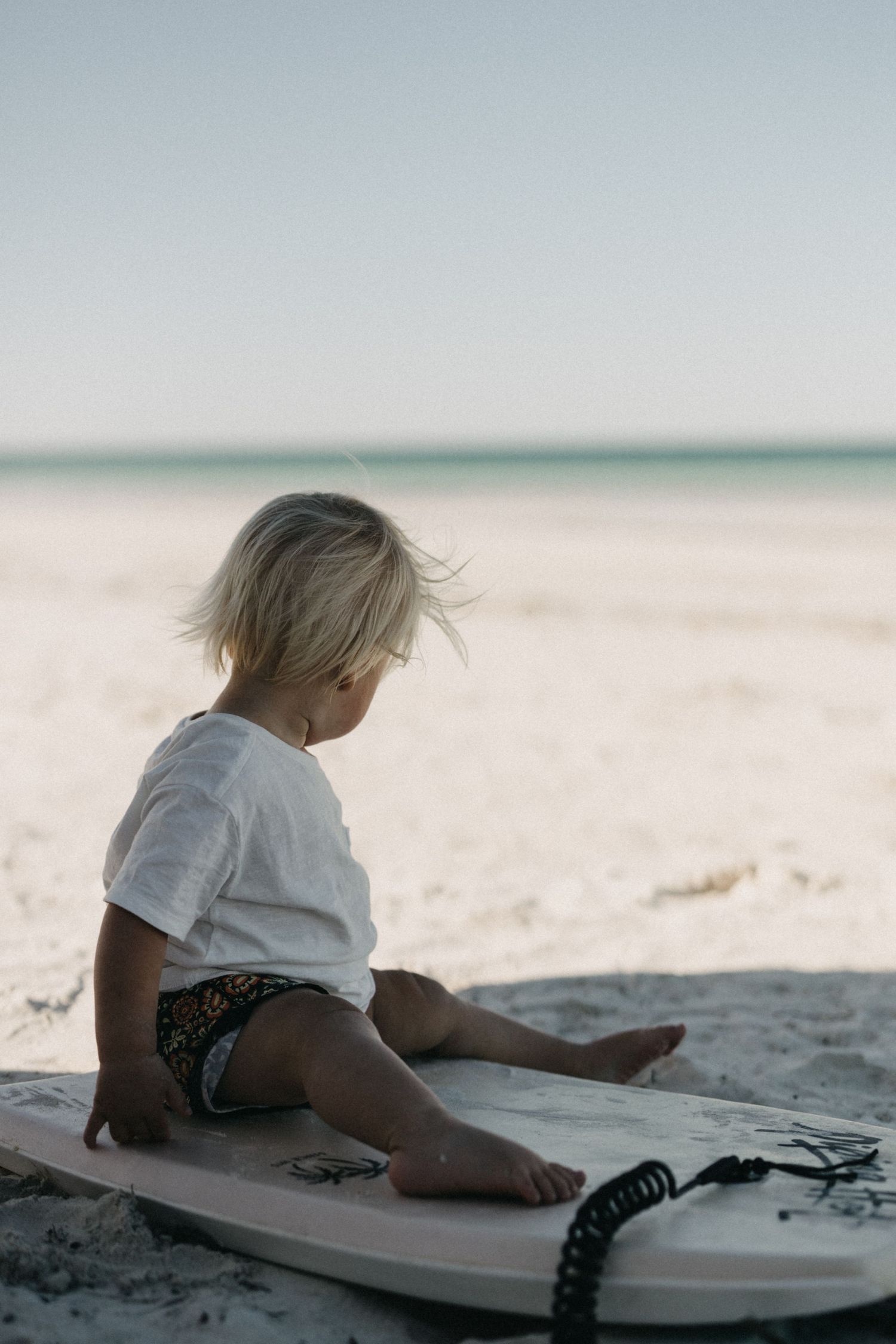 Small child sitting on surfboard on white sandy beach.