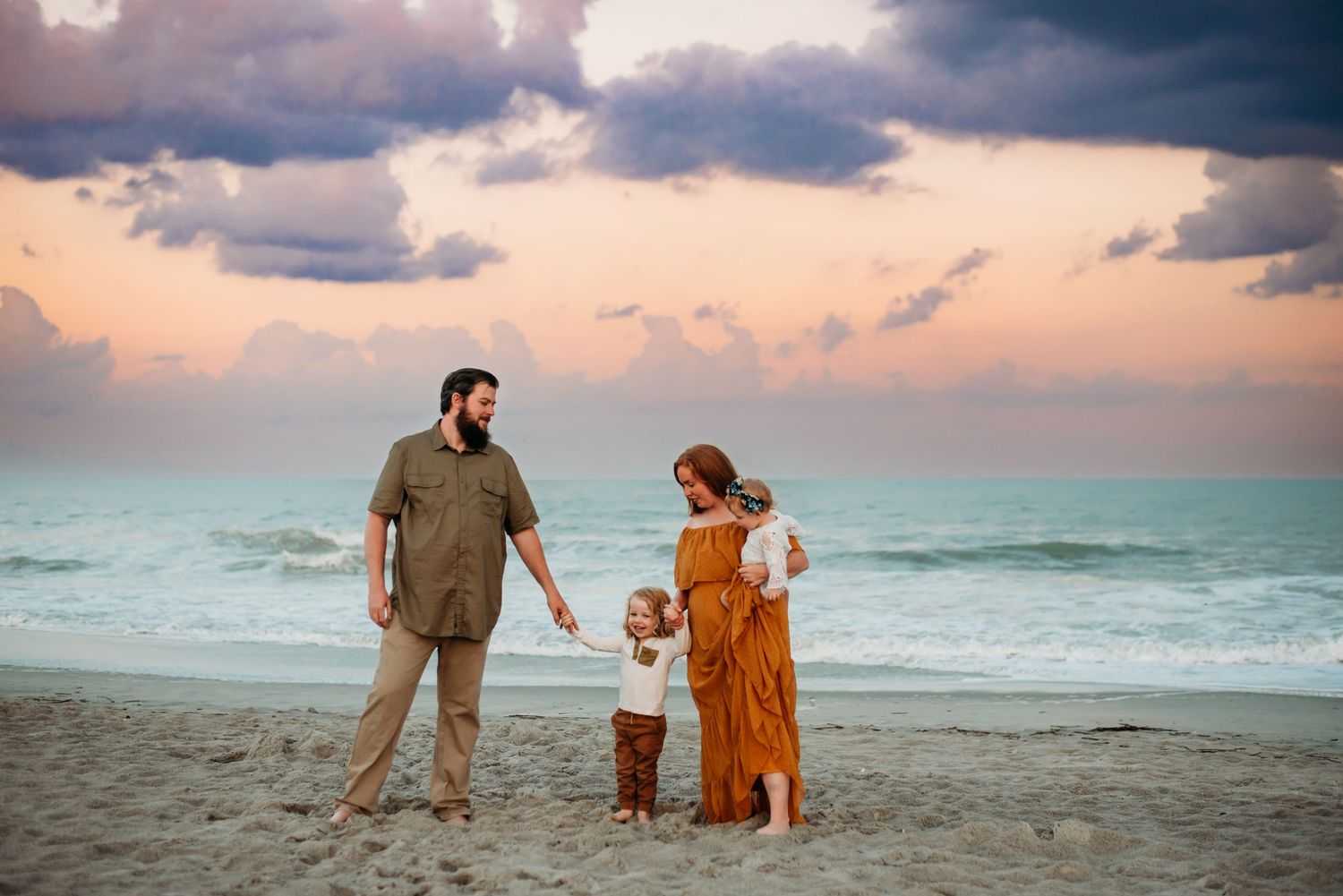 Family of four holding hands on a sandy beach at sunset with dramatic cloudy sky.