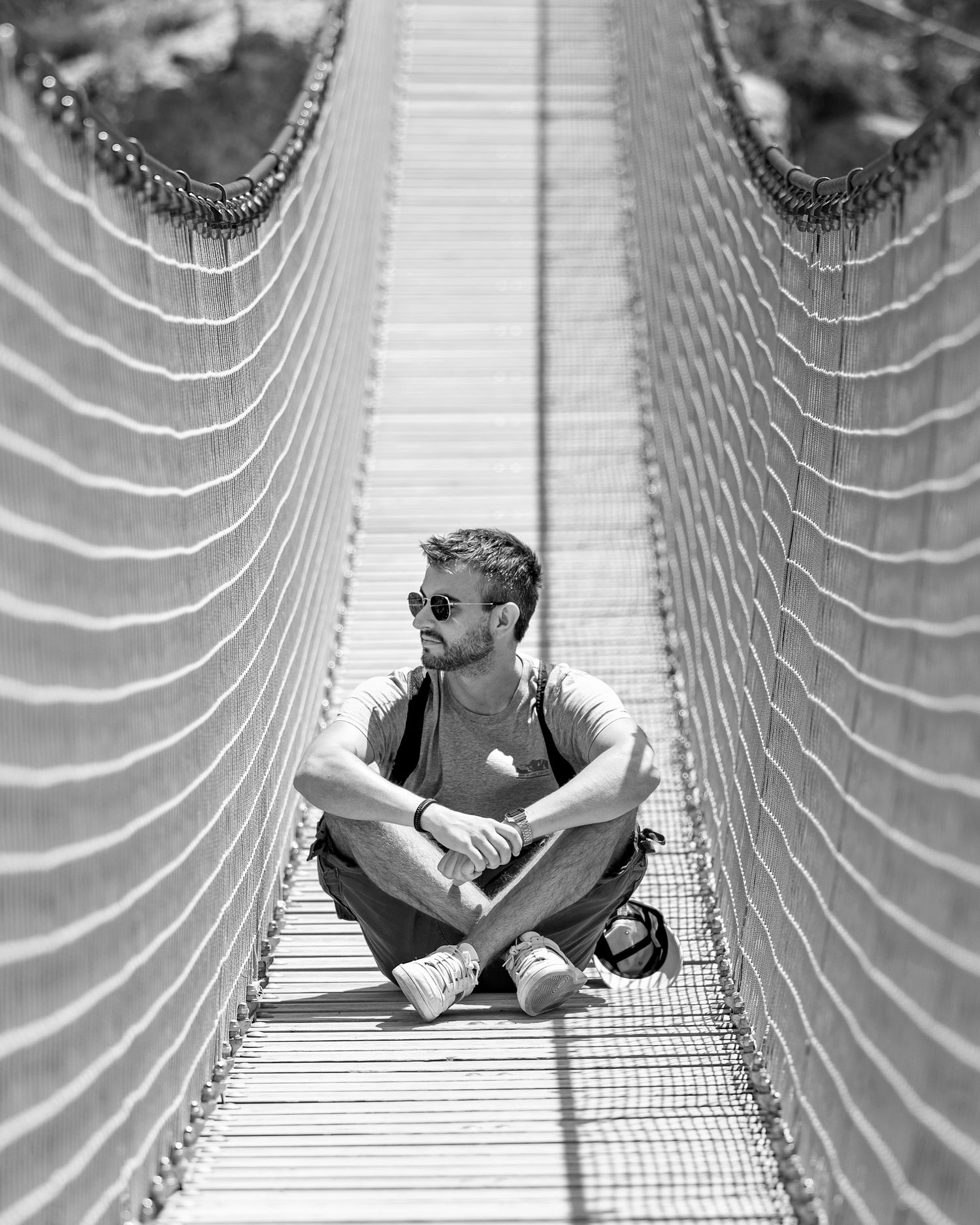 Person sitting cross-legged on suspended rope bridge in dramatic black and white photography.