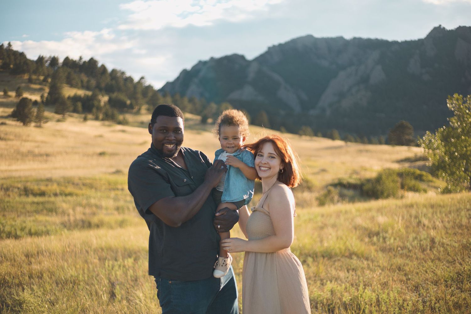 biracial parents posing for camera holding toddler baby boy mountains golden field