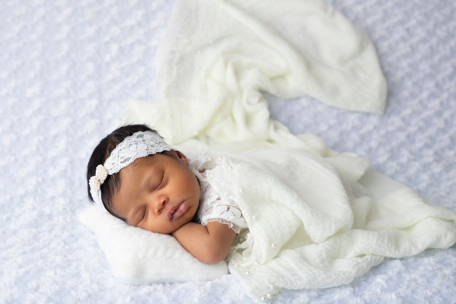 A sleeping newborn in white lace outfit with headband against textured white background.