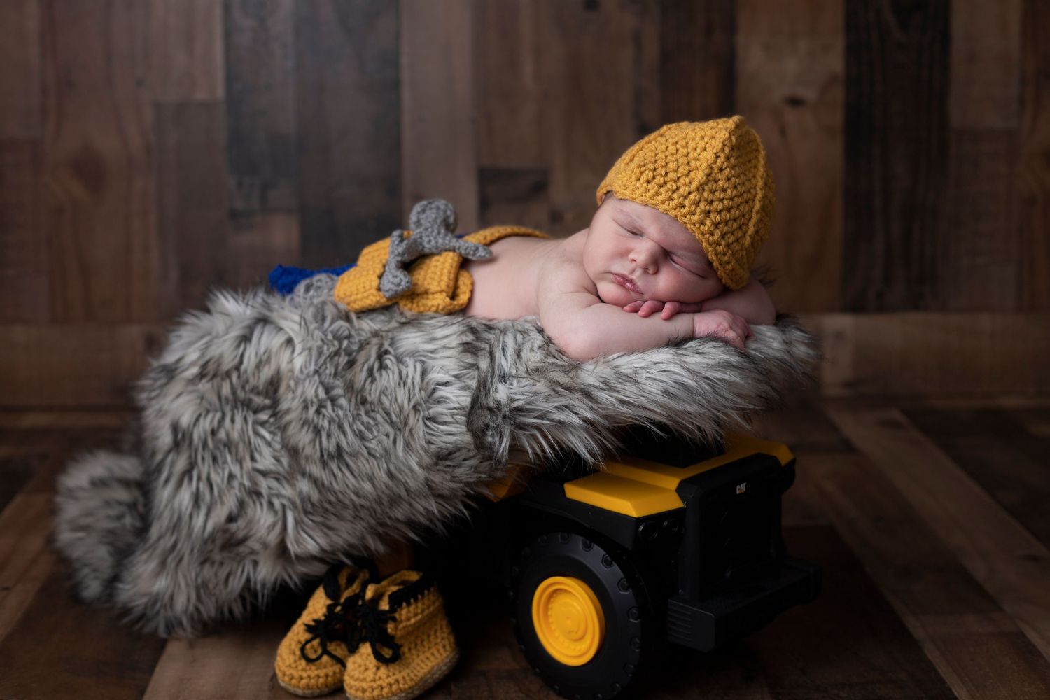 Baby resting on grey fur blanket wearing yellow knit hat in studio portrait.