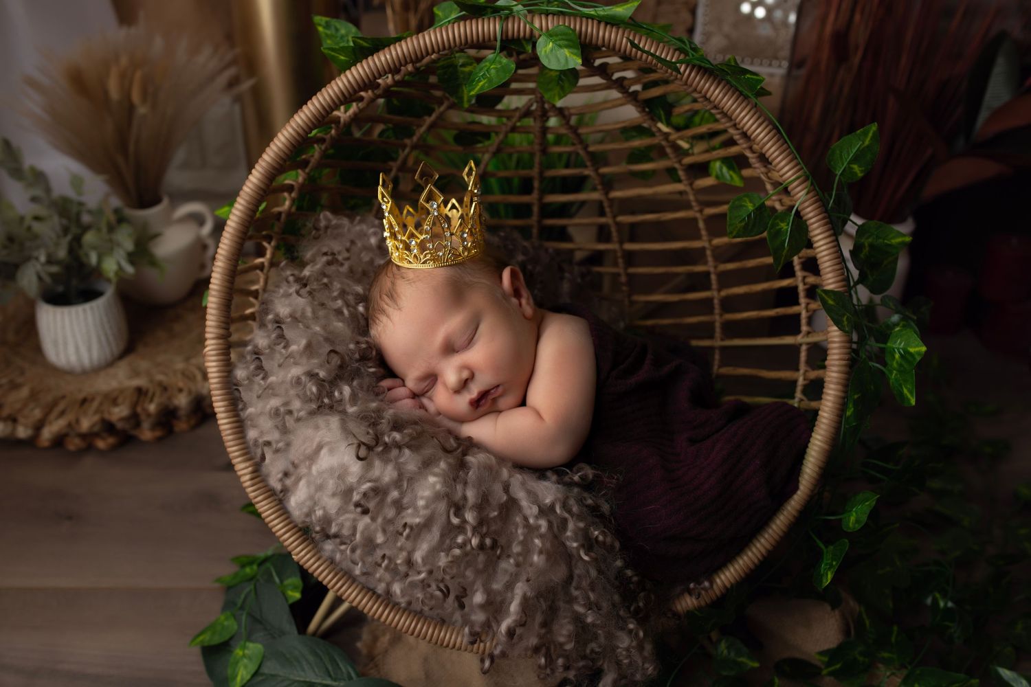 Peaceful newborn sleeping in round rattan chair with tiny crown and greenery.