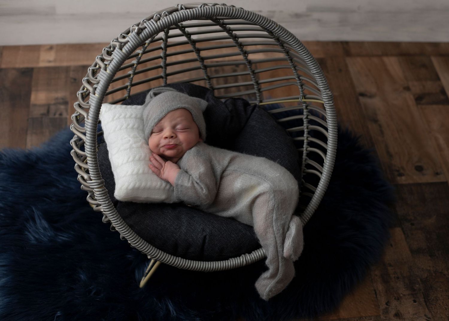 A sleeping baby in grey outfit resting in a round wicker chair on navy blue rug.