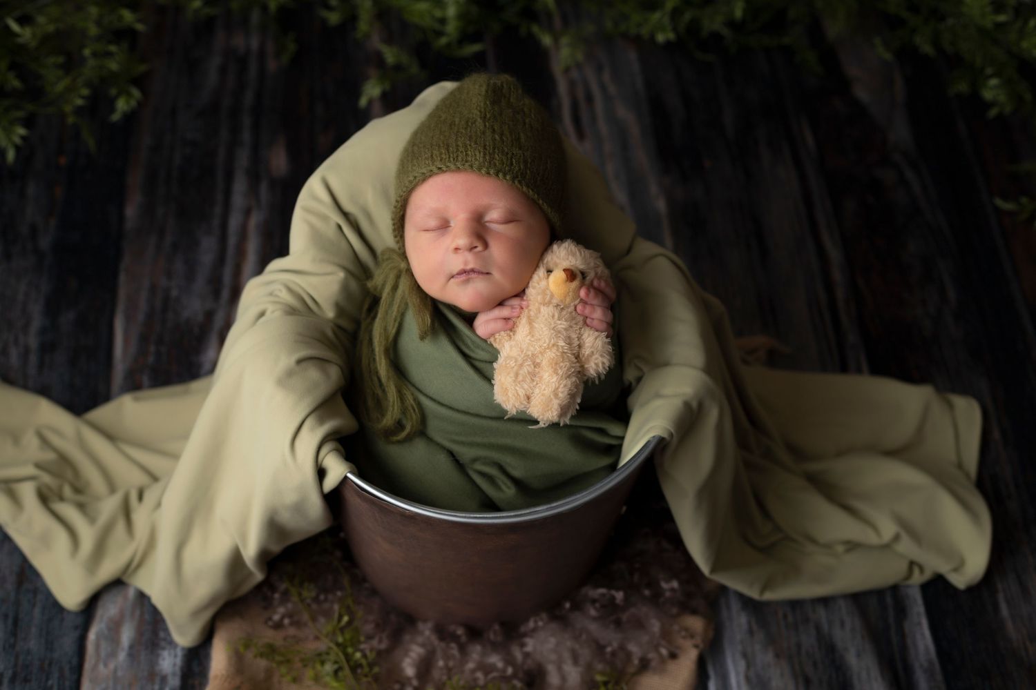 A sleeping newborn wrapped in olive green blankets nestled in a dark metal bowl against a black background.