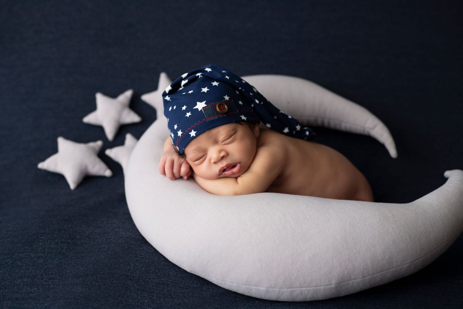 Sleeping newborn on crescent moon prop with stars against dark background.