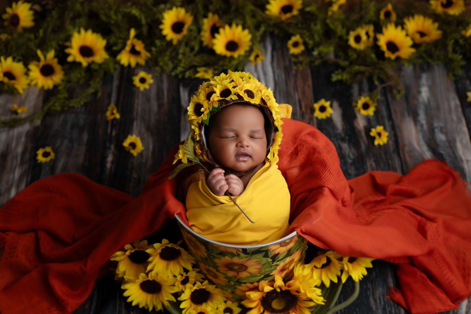 Artistic newborn portrait with vibrant sunflowers and red fabric backdrop.