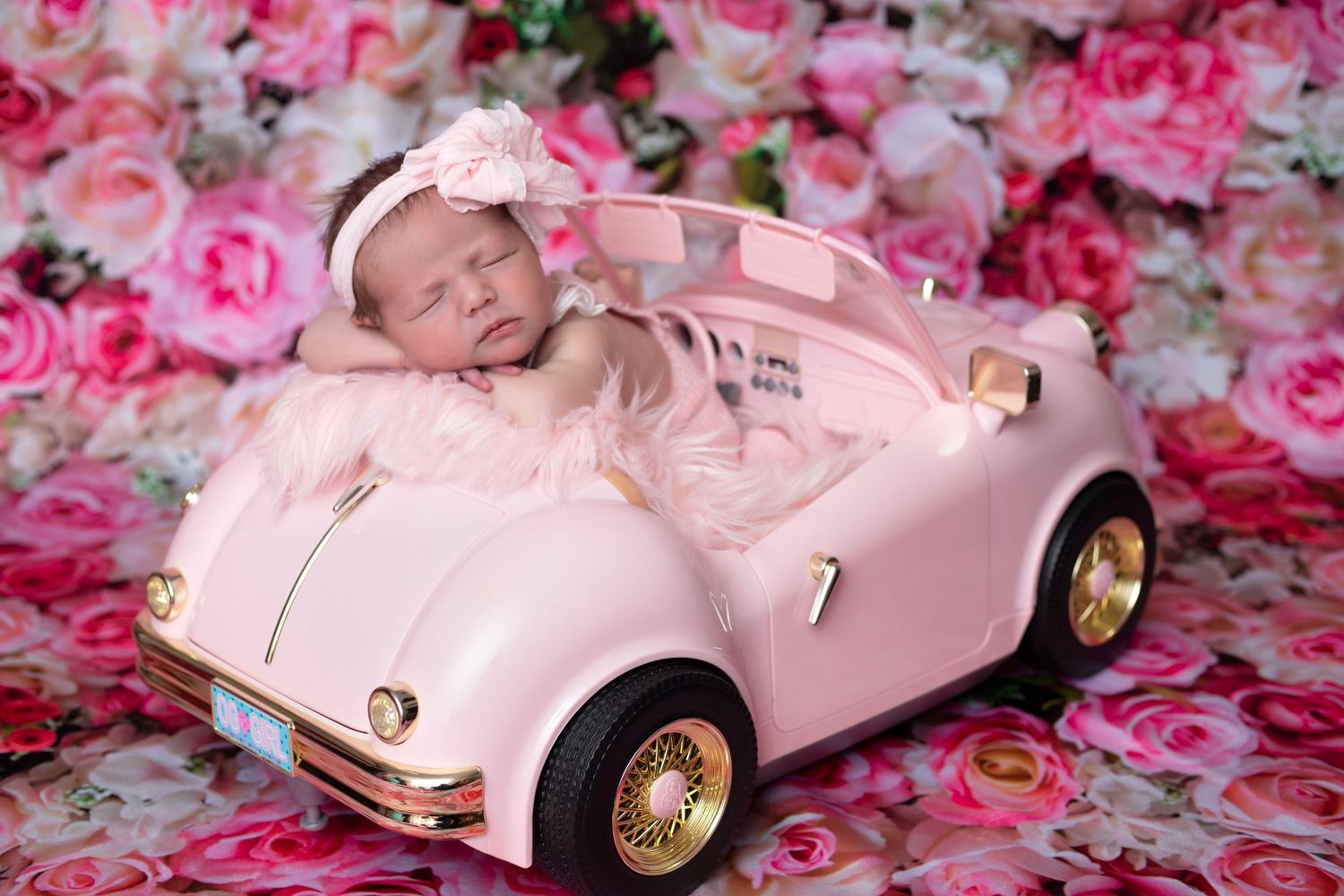 A sleeping baby in a pink vintage toy car surrounded by pink roses.