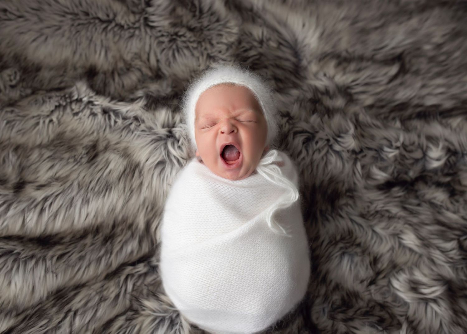 Newborn baby wrapped in white swaddle on grey fur background.