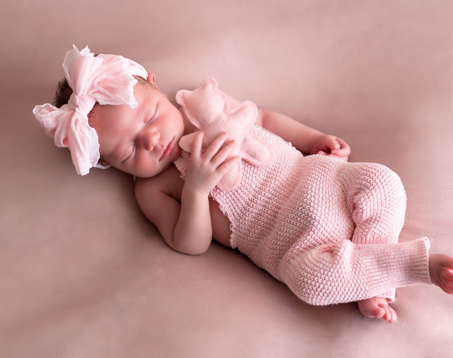 A sleeping newborn wearing pink knit clothing and bow on soft pink background.