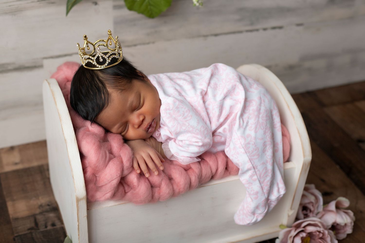 A sleeping baby wearing crown in pink outfit resting in white curved container.