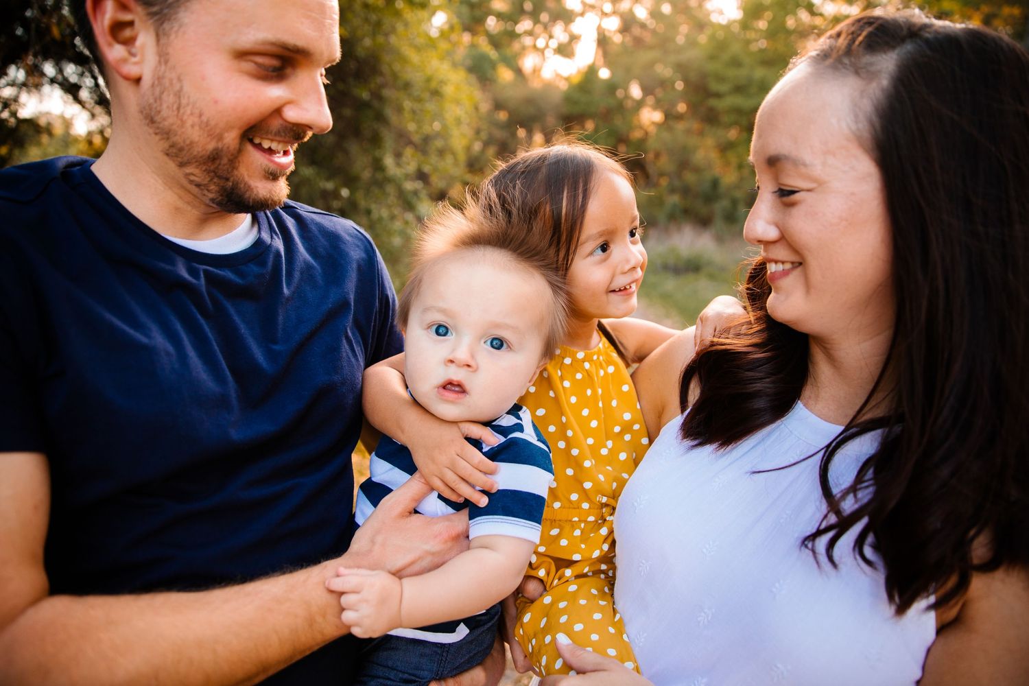 Happy young family with smiling parents holding adorable baby and toddler girl outdoors in golden light.