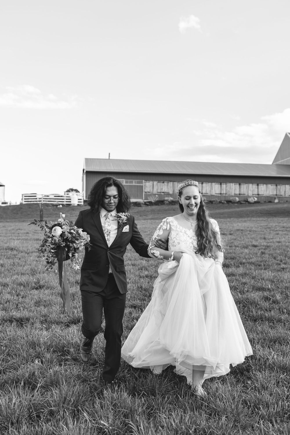 Newlywed couple walking hand in hand across a grassy field with a barn in the background.