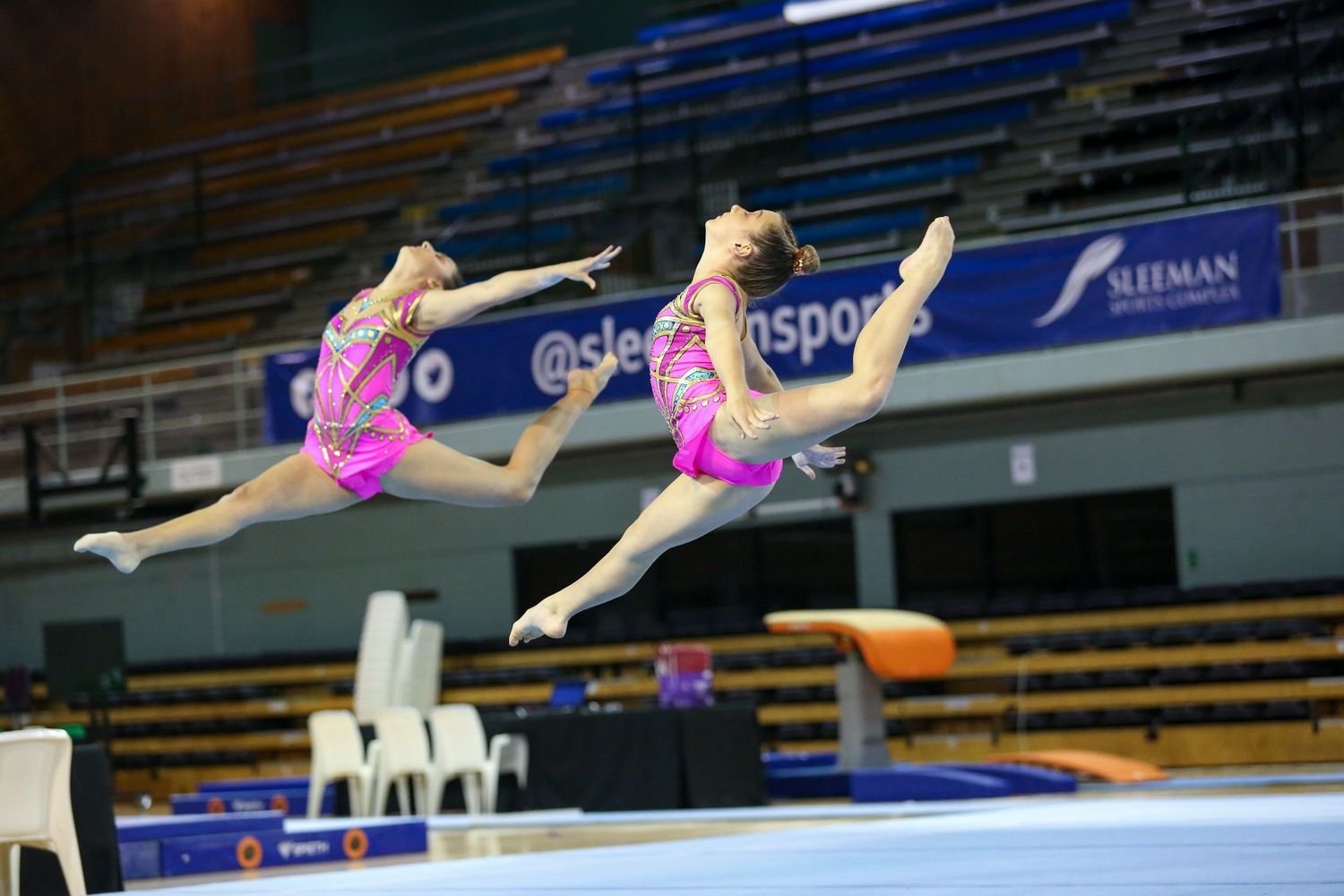 Two gymnasts in pink leotards performing synchronized leap movements during competition.