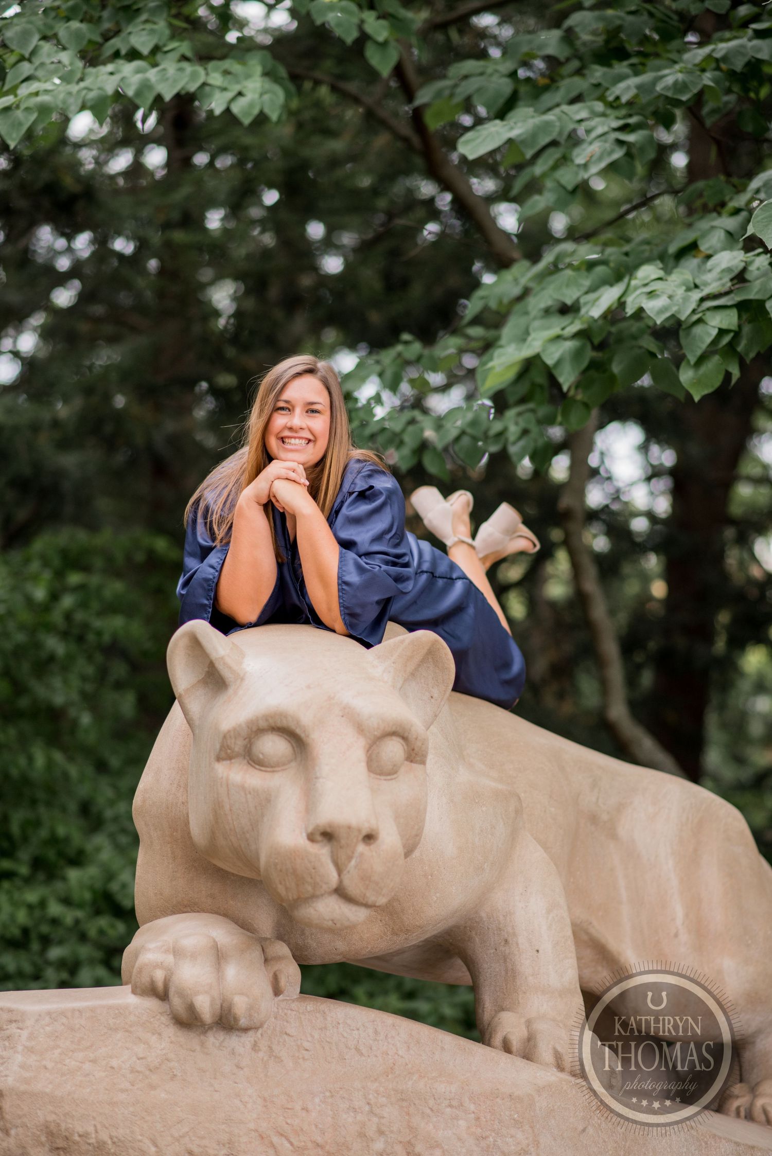 Graduate in navy gown reclines on Penn State's iconic limestone lion sculpture.