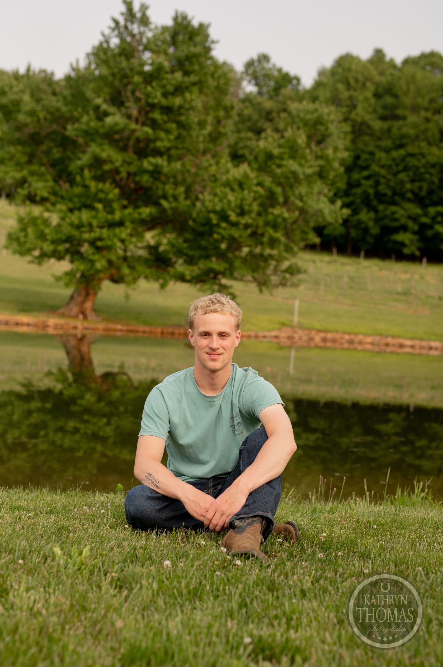 Person sits cross-legged on grassy hill overlooking serene pond landscape.