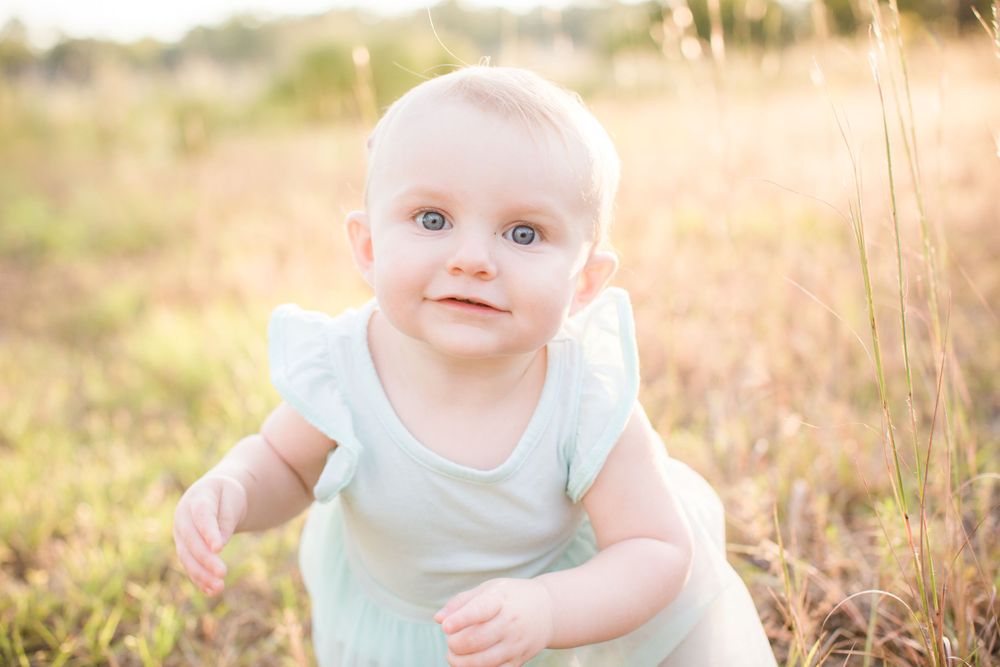 A baby in a white dress sits in golden sunlit grass during sunset.