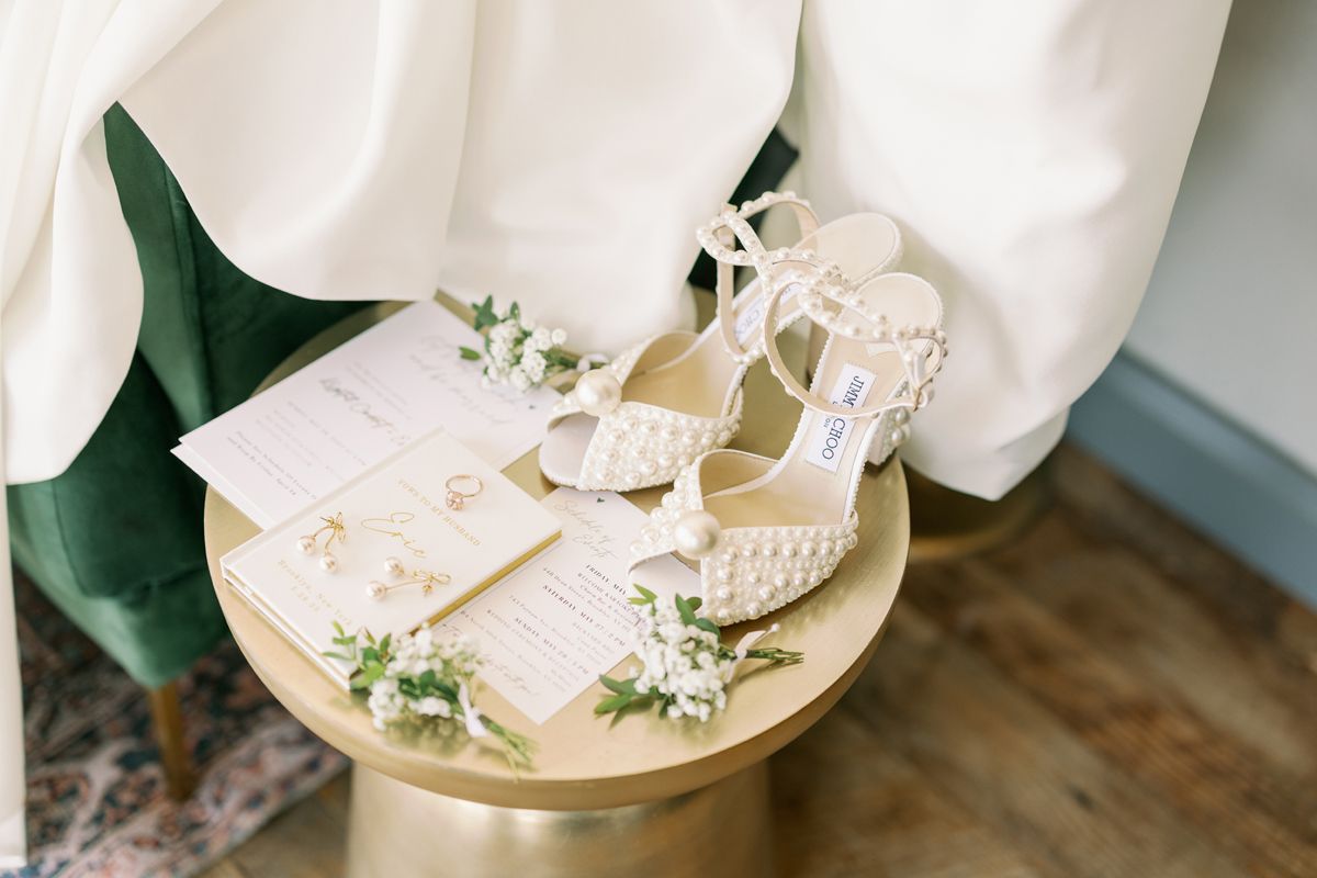 Wedding accessories including white heels and invitations arranged on gold side table.