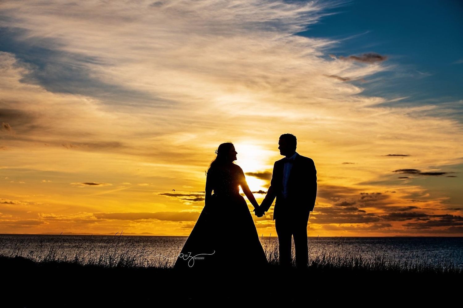 Silhouette of a bride and groom against a dramatic sunset sky by the water.
