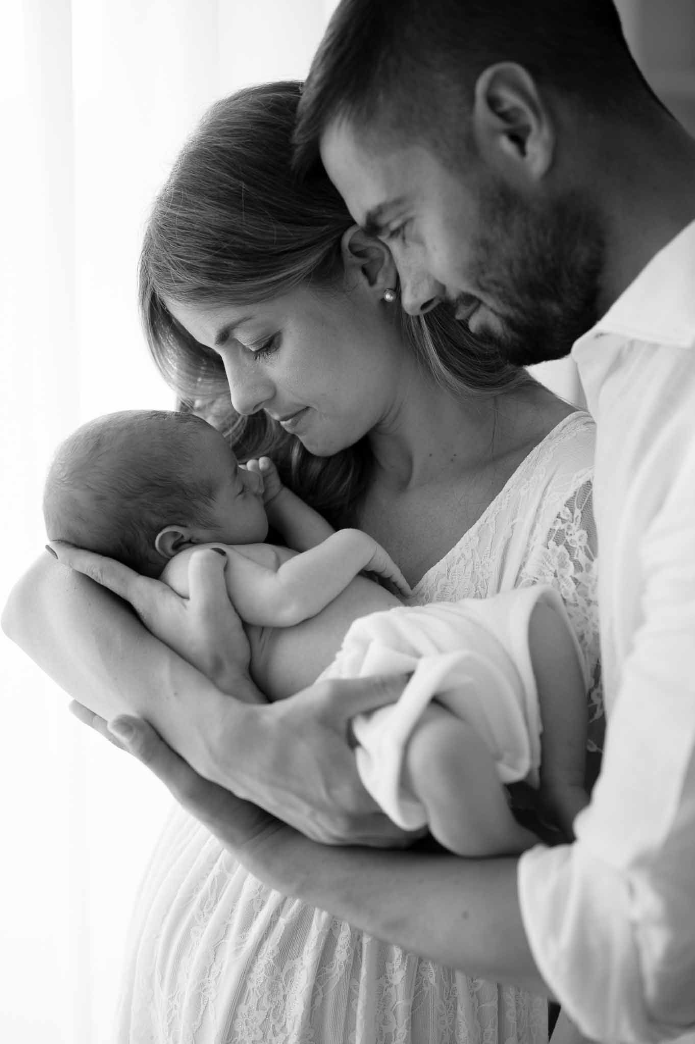 Black and white family portrait showing intimate moment with newborn baby by window.