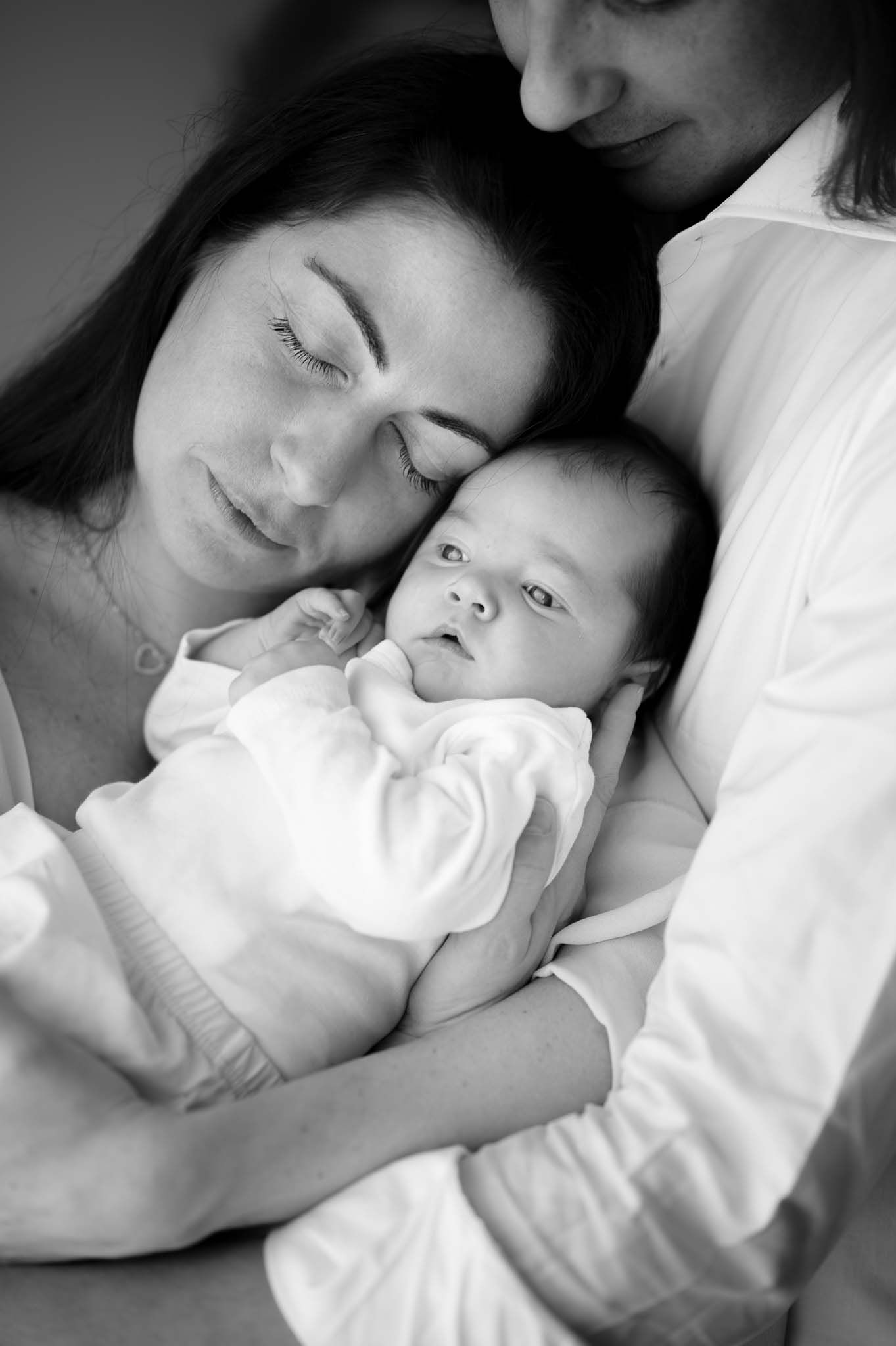 Intimate black and white portrait of parents cradling newborn baby in white clothing.