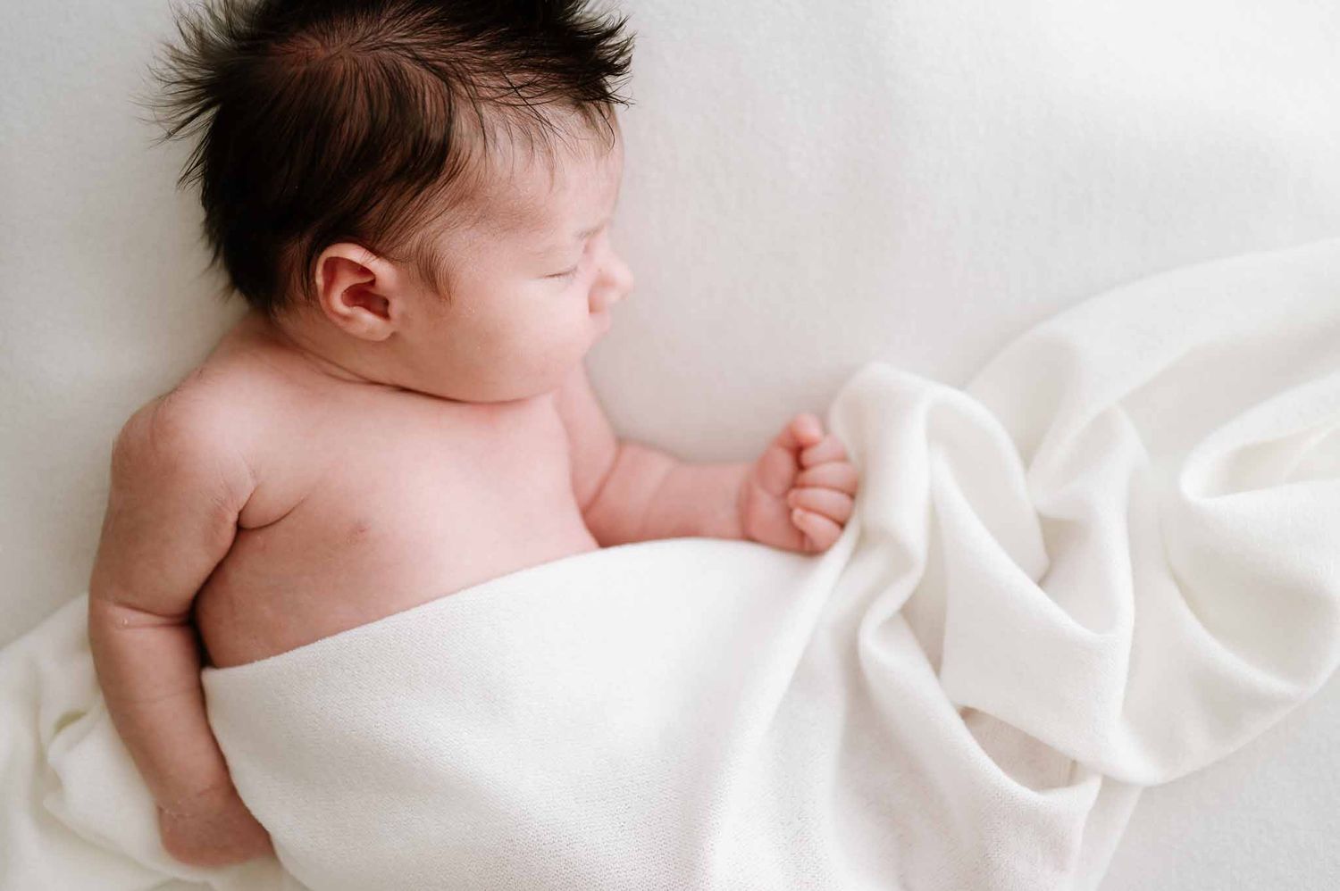 Peaceful sleeping newborn wrapped in soft white blanket on light background.