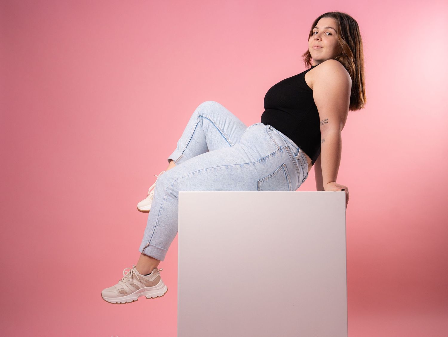 Woman in casual outfit sitting on white cube against pink background.