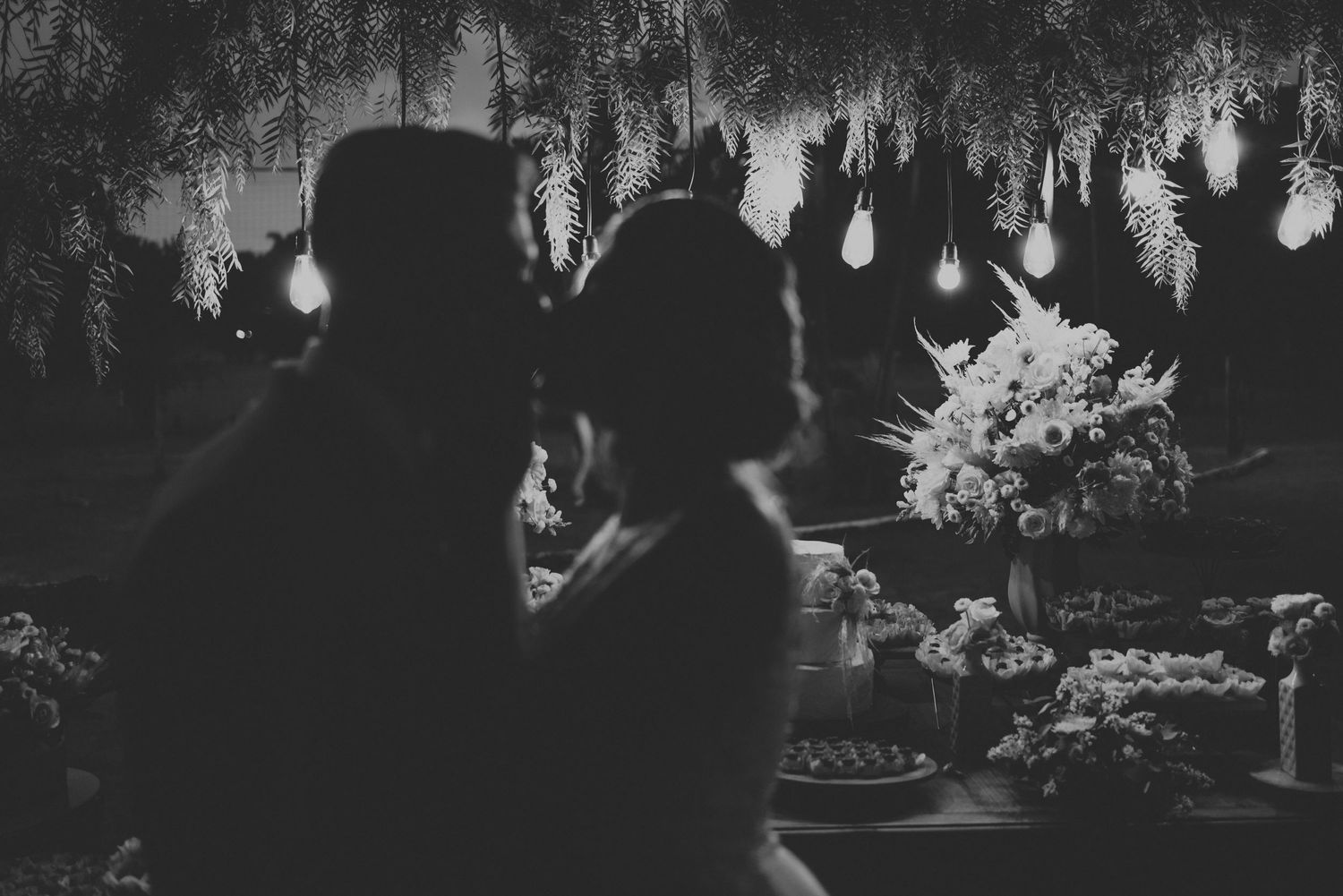 silhueta de noivos em momento intimo durante a festa, com luzes decorativas suspensas (fairy lights) e arranjos florais ao fundo