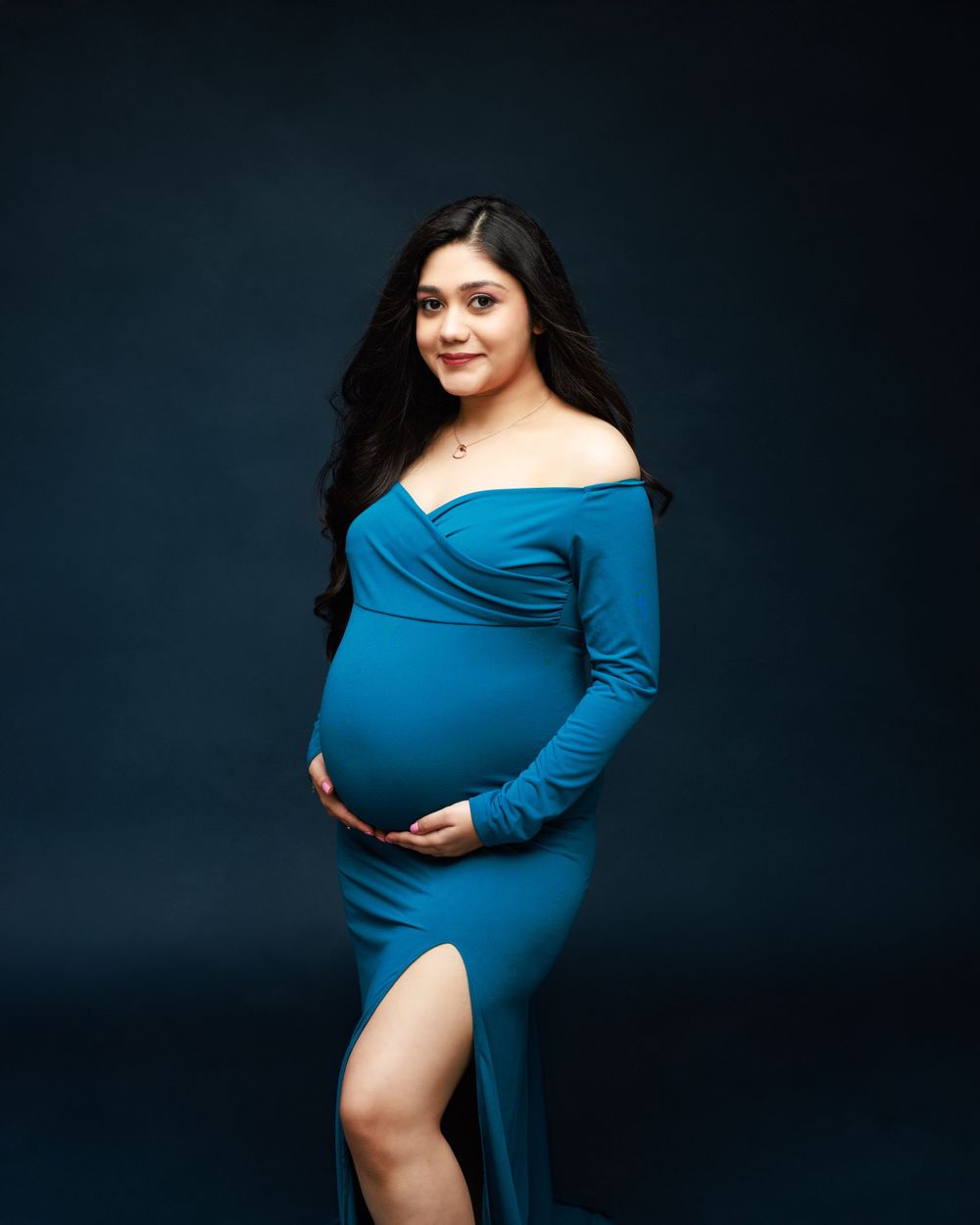 Studio maternity photo featuring blue off-shoulder dress with leg slit against dark backdrop.