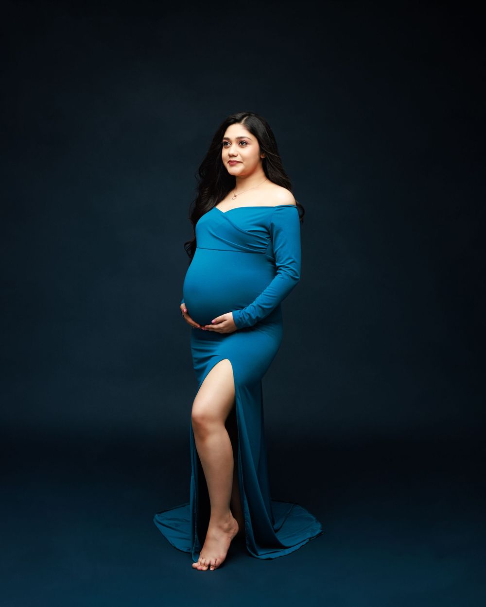 Elegant maternity portrait in fitted blue off-shoulder gown against dark background.